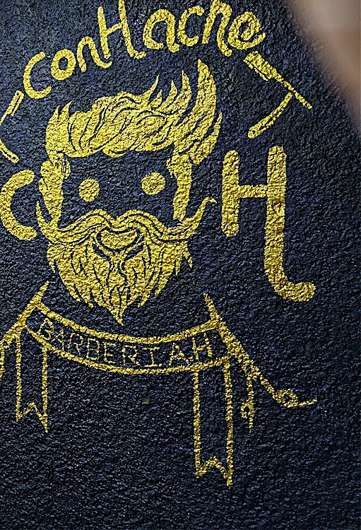 Mural pintado a mano en dorado sobre muro negro: el personaje de ConHache con copete y barba entre una C y una H, con el listón Barbería